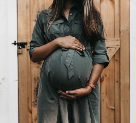 A pregnant woman wearing a dark green dress holding her stomach with both her arms.