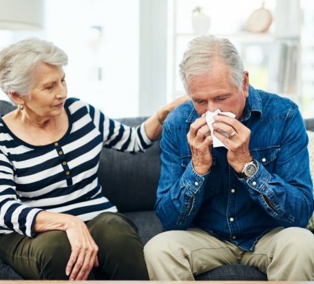 An elderly man is blowing his nose while his wife is comforting him on the sofa.